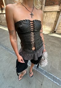 Must musta nahkaineline korsett, millel on ees nöörimisdetailid, koos kihilise halli ja musta riietusega ning sandaalidega.