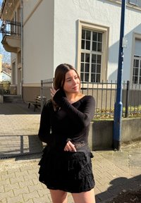 Czarny top z długim rękawem o dopasowanym kroju, zestawiony z czarną mini spódnicą o teksturowanym wykończeniu, z falbankami i kieszeniami. Słoneczne otoczenie na zewnątrz.
