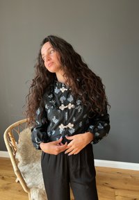 Schwarze Bluse mit blauer Blumenstickerei, hohe Halslinie und puffige Ärmel. Silberne Hardware-Akzente an der Vorderseite.