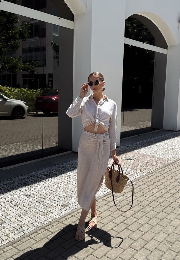 Mujer con crop top atado de color beige claro y falda midi, sosteniendo un bolso tejido, ajustándose las gafas de sol mientras camina por una acera soleada.