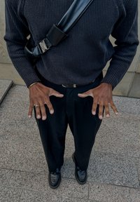 Maglione a coste nero, pantaloni neri e scarpe nere. Mani posizionate in avanti, mostrando gli anelli. Fibbia riflettente sulla tracolla.