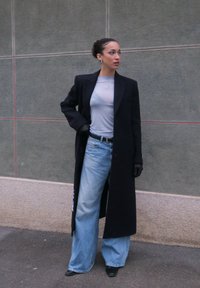 Femme avec chignon portant un long manteau noir, un haut gris clair, un jean bleu à jambes larges, des gants noirs et des chaussures noires à bouts pointus, debout près d'un mur gris.