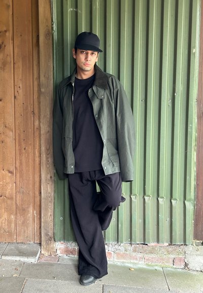 Camiseta negra debajo de una camisa overshirt verde oliva con cierre de botones. Pantalones negros holgados y una gorra negra ajustada. Fondo de ladrillo y metal.