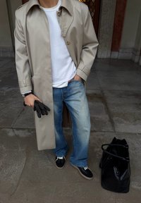 Trench-coat beige, chemise blanche, jean bleu clair, baskets noires, tenant des gants noirs. Sac cabas noir à côté. Surface en pierre texturée en dessous.
