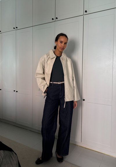 Chaqueta de cuero brillante color crema, blusa ajustada oscura, pantalones de mezclilla de pierna ancha de tiro alto en oscuro, bailarinas negras simples, contra un fondo de armario blanco.