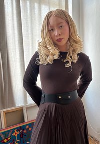 Vestido castanho de mangas longas com saia plissada e cinto largo preto com ferragens douradas. Cabelo loiro longo e estilizado. Iluminação suave e natural.