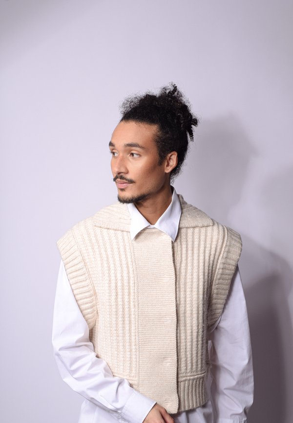 Gilet lavorato a maglia di colore crema con texture a coste e spalline larghe, indossato sopra una camicia bianca, caratterizzato da un colletto alto distintivo e una tasca frontale.