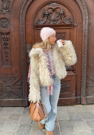 Mujer con abrigo de piel sintética beige, gorro rosa y vaqueros claros, sosteniendo una taza de café y un bolso marrón, de pie frente a una puerta de madera ornamentada.