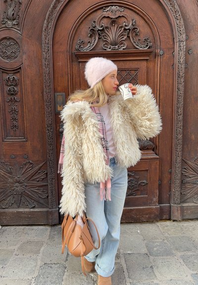 Mujer con abrigo de piel sintética beige, gorro rosa y vaqueros claros, sosteniendo una taza de café y un bolso marrón, de pie frente a una puerta de madera ornamentada.
