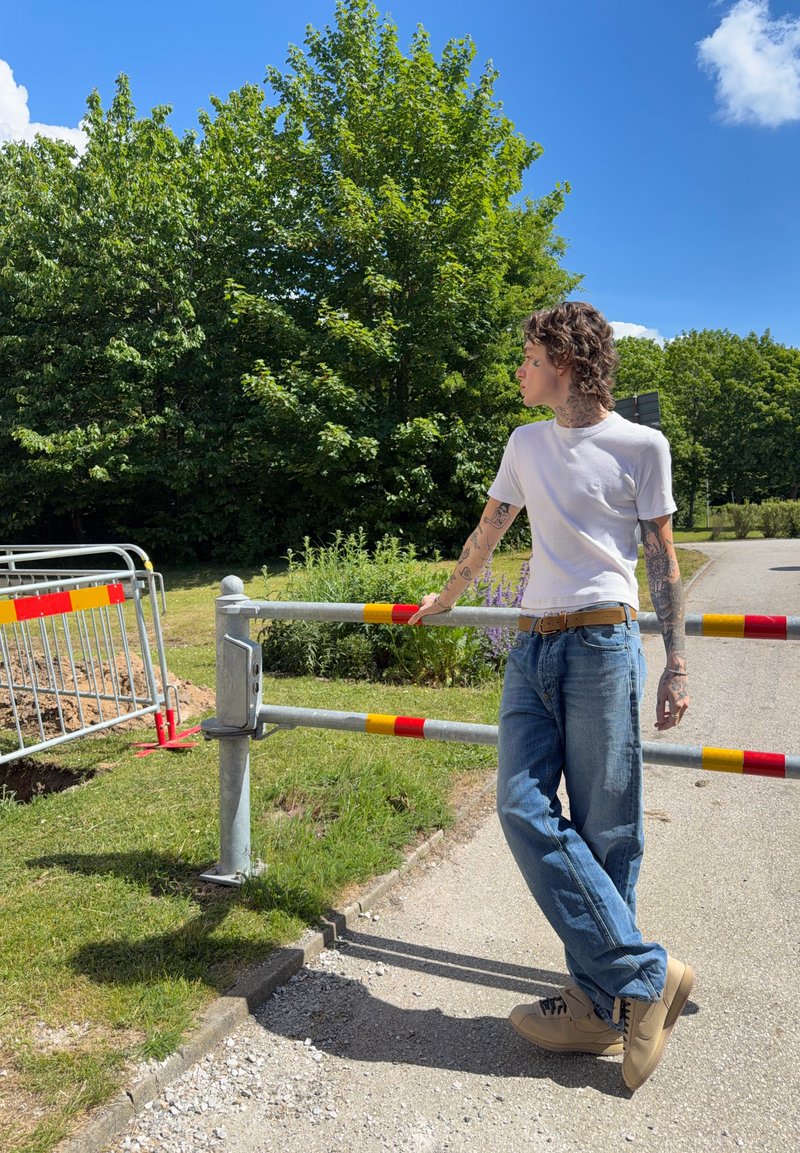 Jeune personne avec des tatouages portant un t-shirt blanc et un jean, appuyée contre une barrière métallique sur un chemin ensoleillé avec des arbres et un ciel bleu derrière.