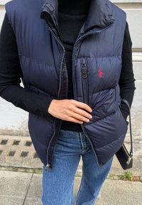 Tumesinine puffer-vest kõrge krae ja tõmblukuga, millel on väike punane logo, kantud musta topi ja siniste teksade peal.