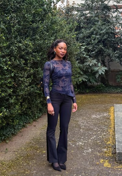 Mujer de pie al aire libre con una blusa oscura translúcida con flores, pantalones negros acampanados y zapatos puntiagudos, con arbustos verdes al fondo.