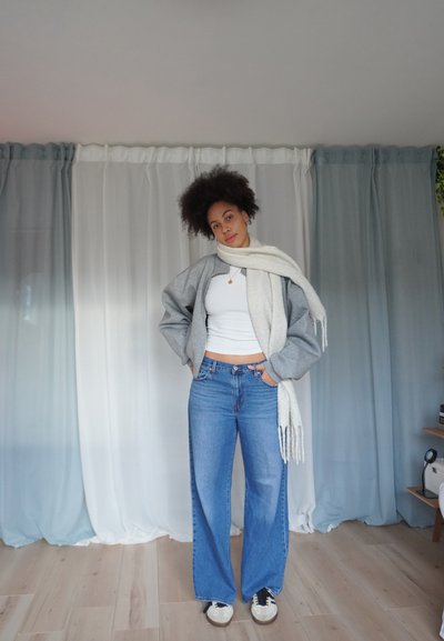 Chaqueta bomber gris, camiseta sin mangas blanca, vaqueros de pierna ancha azul y una bufanda de color claro. Ambientado contra cortinas drapeadas blancas y azules.