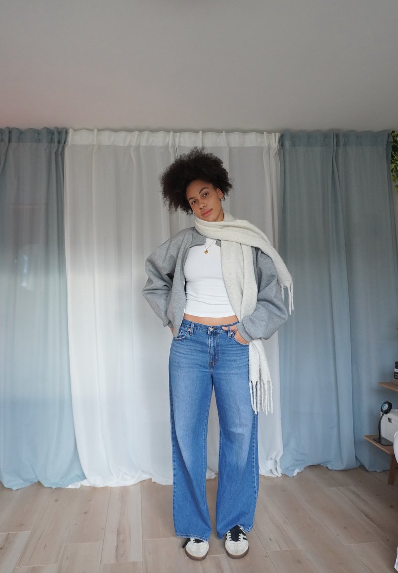 Giacca bomber grigia, canottiera bianca, jeans blu a gamba larga e una sciarpa di colore chiaro. Sullo sfondo tendaggi bianchi e blu.