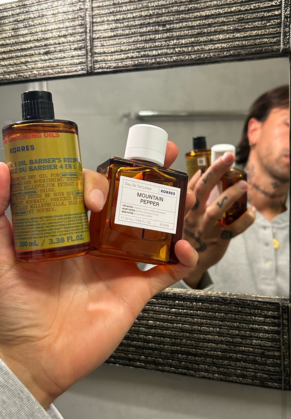 Egy kéz két borostyánszínű üveget tart: Korres 4 az 1-ben Borbély recept olaj és Mountain Pepper Eau De Toilette, a fürdőszoba tükörében tükröződve.