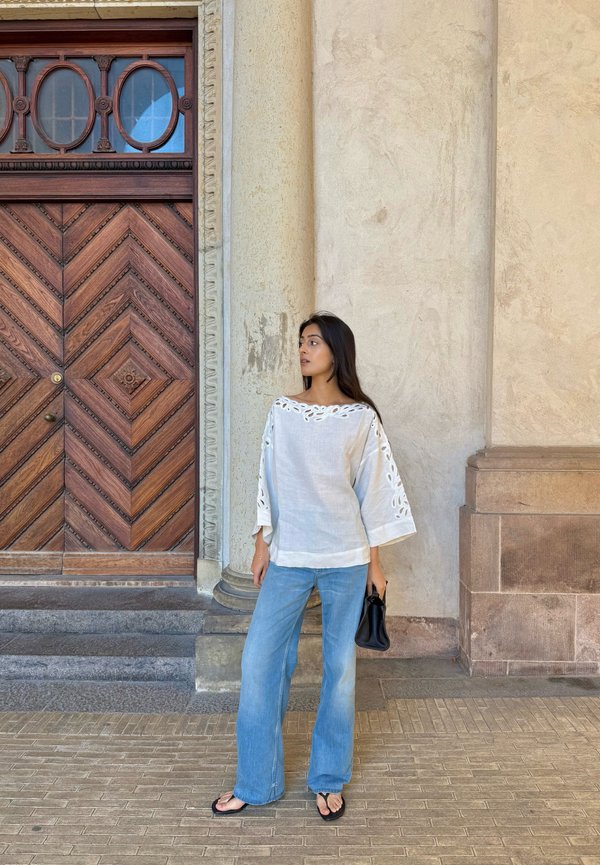 Vrouw in een witte blouse en blauwe spijkerbroek met een zwarte handtas staat bij een sierlijke houten deur en een stenen zuil in een stedelijke omgeving.
