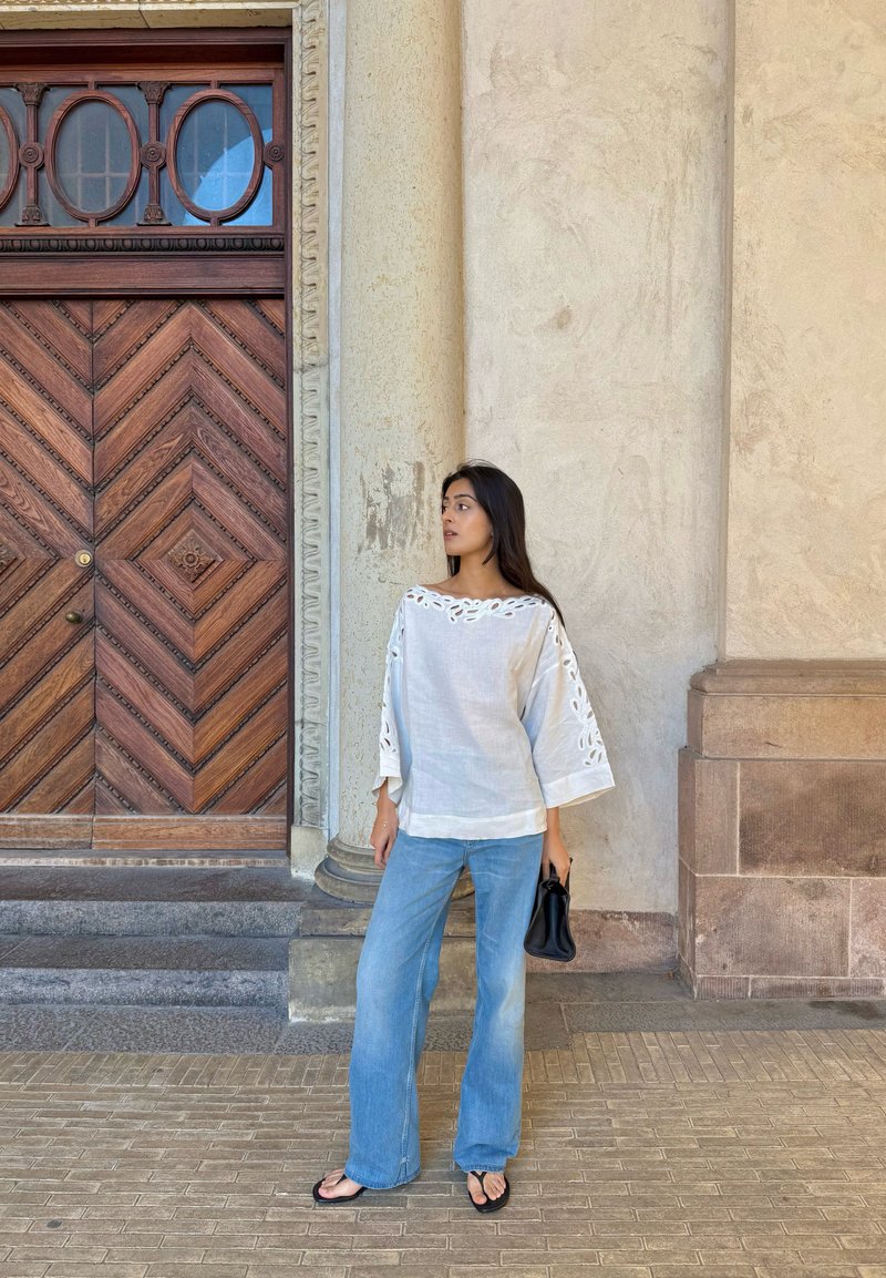 Femme en blouse blanche et jean bleu tenant un sac noir se tient près d'une porte en bois ornée et d'une colonne en pierre dans un cadre urbain.