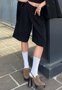 Crne hlače s naborima, uparene s bijelim rebrastim čarapama i smeškim suknjicama od antilopa s potpeticama. Model nosi crni blejzer i stoji na otvorenom.