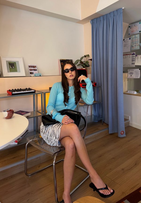 Femme portant des lunettes de soleil, un cardigan bleu et une jupe à imprimé zèbre, assise sur une chaise transparente dans une pièce avec un plancher en bois et des étagères.