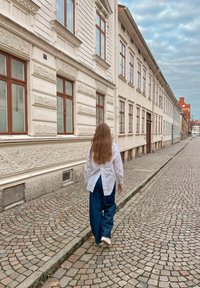 Voľne padnutá biela košeľa a tmavomodré široké nohavice, kráčajúca po dláždenej ulici vedľa textúrovaných bielych budov s drevenými oknami.