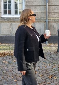Juodas vilnos mišinio švarkas su sidabriniais mygtukais ir priekinėmis kišenėmis, vilkimas ant baltos marškinių. Asmuo laiko kavos puodelį.