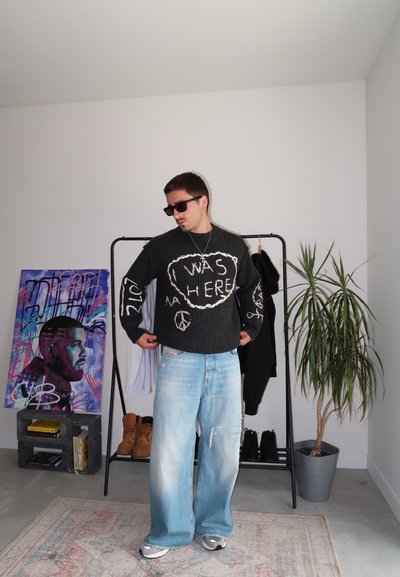 Suéter gris con diseño de texto blanco, jeans azules holgados y gafas de sol. Conjunto casual con detalles gráficos y un ajuste relajado. Planta y arte al fondo.