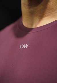 Κοντινό πλάνο ενός ατόμου που φοράει μια μπορντό μπλούζα με λαιμόκοψη, με το γράμμα "ICIW" τυπωμένο σε λευκό στο στήθος.