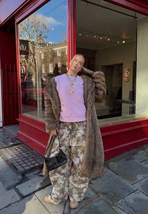 Persona con abrigo largo de piel marrón, camisa rosa, pantalones de camuflaje, zapatos beige, sosteniendo un bolso negro, de pie junto a una vitrina de tienda con marco rojo en la acera de la ciudad.