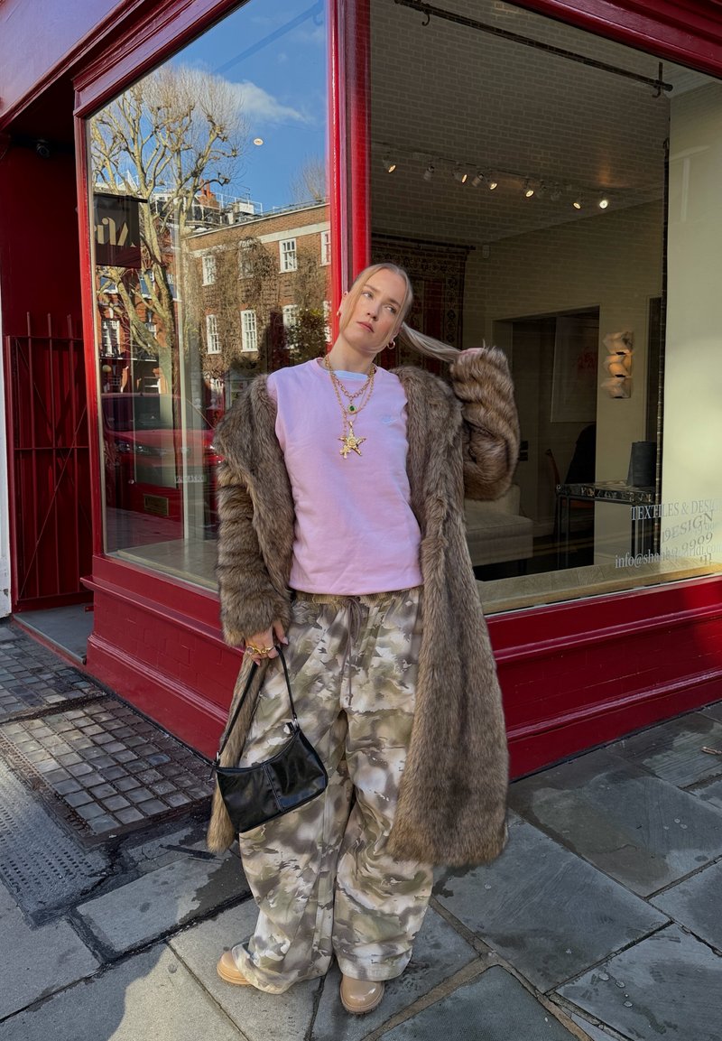 Personne portant un long manteau en fourrure marron, une chemise rose, un pantalon camouflage, des chaussures beiges, tenant un sac à main noir, debout près d'une vitrine de magasin encadrée en rouge sur le trottoir de la ville.