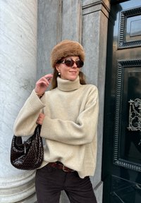 Beiger Strickturtleneck-Pullover mit weiten Ärmeln, kombiniert mit dunklen Cordhosen. Brauner Pelzhut, Sonnenbrille und eine geflochtene braune Handtasche.