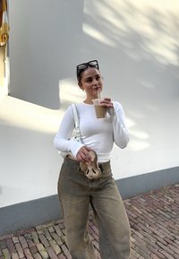 Hvit langermet skjorte, høy midje khaki bukser, holder en klar kopp med en brun drink og en tan veske som ligner på en sko.