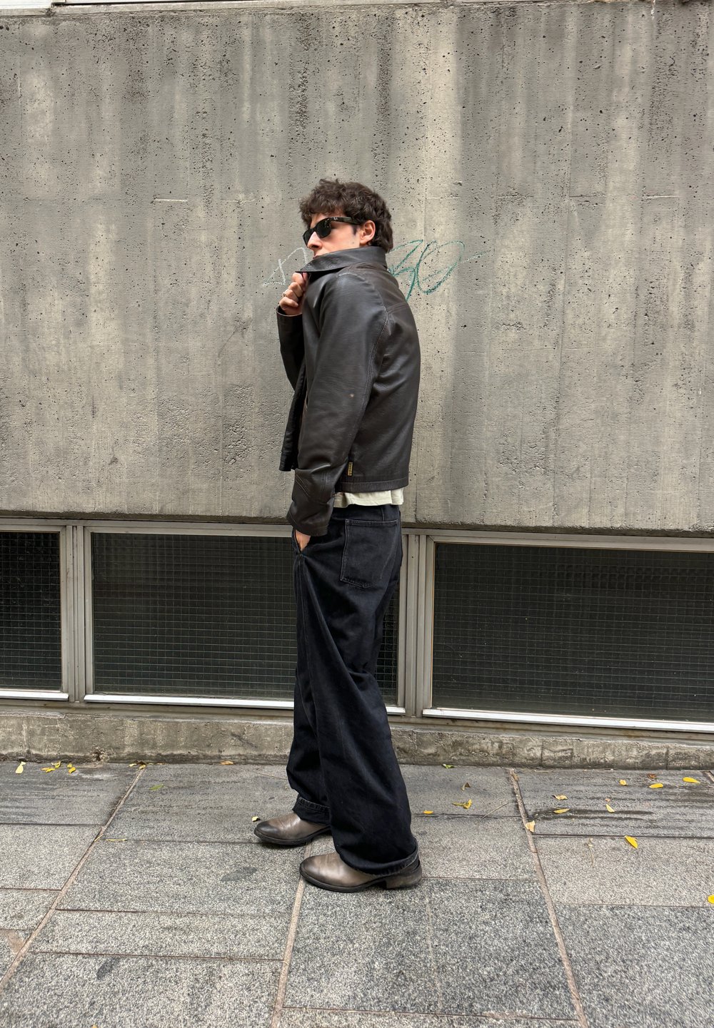 Chaqueta de cuero negra, camisa blanca, pantalones anchos negros y botas grises. De pie contra una pared de hormigón texturizada con grandes ventanales en la parte inferior.
