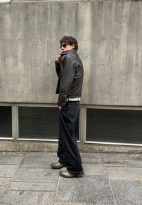 Chaqueta de cuero negra, camisa blanca, pantalones anchos negros y botas grises. De pie contra una pared de hormigón texturizada con grandes ventanales en la parte inferior.