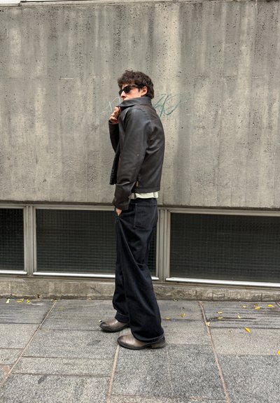 Chaqueta de cuero negra, camisa blanca, pantalones anchos negros y botas grises. De pie contra una pared de hormigón texturizada con grandes ventanales en la parte inferior.