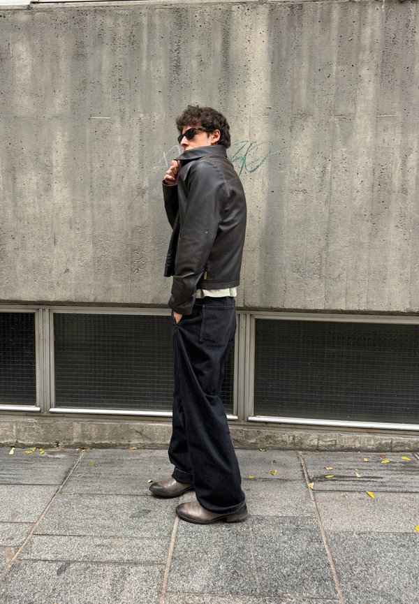 Chaqueta de cuero negra, camisa blanca, pantalones anchos negros y botas grises. De pie contra una pared de hormigón texturizada con grandes ventanales en la parte inferior.