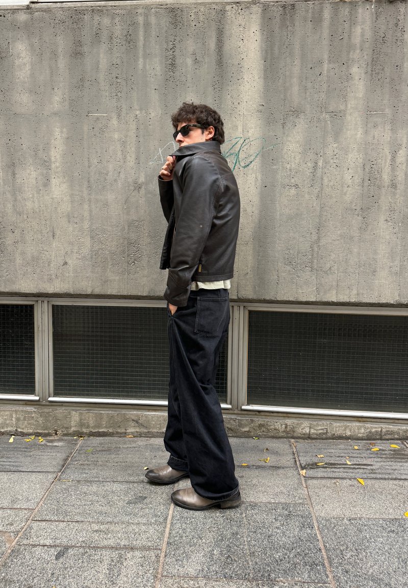 Veste en cuir noire, chemise blanche, pantalon noir ample et bottes grises. Debout contre un mur en béton texturé avec de grandes fenêtres en dessous.