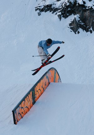 Esquiador con chaqueta azul y casco realiza un truco en el aire sobre una barra cubierta de nieve con grafiti naranja en una pendiente nevada de la montaña.