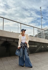 Biały top bez rękawów, kremowa marynarka, oversize'owe dżinsy, a także koszula w kratę związana w talii. Niebieska torebka i dżinsowa czapka dopełniają całość.