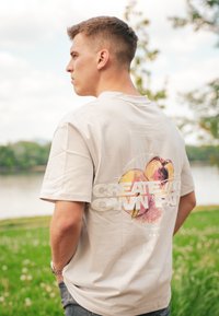 Μπεζ βαμβακερό T-shirt με πολύχρωμο γραφικό σχέδιο στην πλάτη που απεικονίζει το κείμενο "ΔΗΜΙΟΥΡΓΗΣΤΕ ΤΟΝ ΔΙΚΟ ΣΑΣ ΔΡΟΜΟ" με έντονα γράμματα. Κοντά μανίκια.