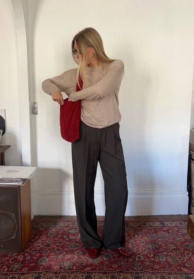 Top de manga larga marrón, pantalones oscuros de cintura alta y bolso de tela rojo. La persona está metiendo la mano en el bolso. Fondo claro.