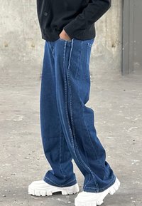 Široke modre jeans hlače z dvema stranskima črtama, z udobnim krojem in obrnjenimi robovi, v kombinaciji z masivnimi belimi platformnimi čevlji.