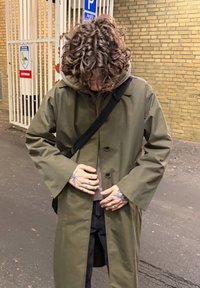 Palton verde măsliniu, rezistent la apă, cu croială lejeră, m�âneci largi și doi nasturi. Purtat peste un hanorac gri și pantaloni negri.