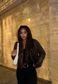 Giacca di pelle marrone, top nero e pantaloni neri. Borsa bianca con una texture liscia. Parete con una finitura in pietra chiara.