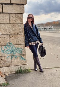 Sort læderjakke over en ternet skjorte, kombineret med marineblå leggings og brune knæhøje støvler. Holder en sort håndtaske og læner sig op ad en stenspiral.