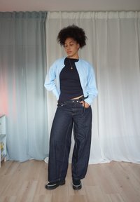 Lichtblauwe cardigan over een zwart topje, gecombineerd met donker denim wijde jeans. De jeans hebben een strikdetail in de taille. Zwarte schoenen.