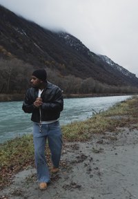 Veste en cuir noire, chemise blanche et jean bleu à jambes larges portés par une personne près d'une rivière, avec des montagnes dans un arrière-plan brumeux.