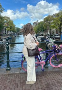 Beige stickad tröja, vita vida byxor och en brun väska. Bakom, en lila cykel med rosa detaljer, blommor och en kanal under en molnig himmel.