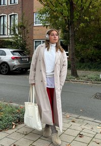 Ljust beige trenchcoat över en vit topp, röda leggings och beige stövlar. Håller en krämfärgad tote-väska. Grå bil och grönska i bakgrunden.