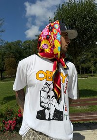 Fehér póló fekete grafikával és sárga 'OBEY' felirattal. Színes virágmintás sál a fej köré kötve. Szabadtéri környezet.