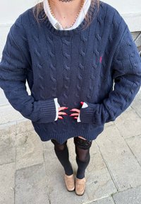 Personne portant un pull marine en maille torsadée, poignets de chemise blancs, bas résille noirs et chaussures beiges, debout sur un pavé en pierre.